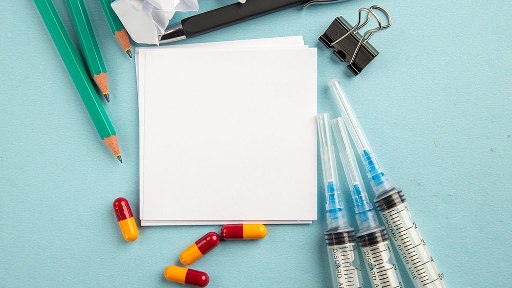 Symbolbild: Blick von oben auf ein Blatt Papier, darum herum Pille, Spritzen und Stifte