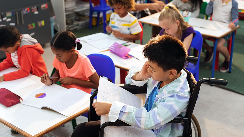 Ein Junge sitzt im Rollstuhl in einem Klassenzimmer und lernt mit seinen Mitschüler:innen