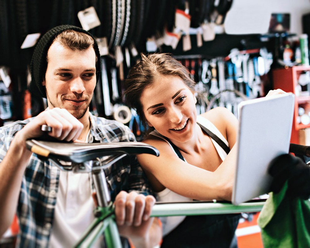 Un jeune homme et une jeune femme réparent un vélo; la femme montre quelque chose à l’homme sur une tablette