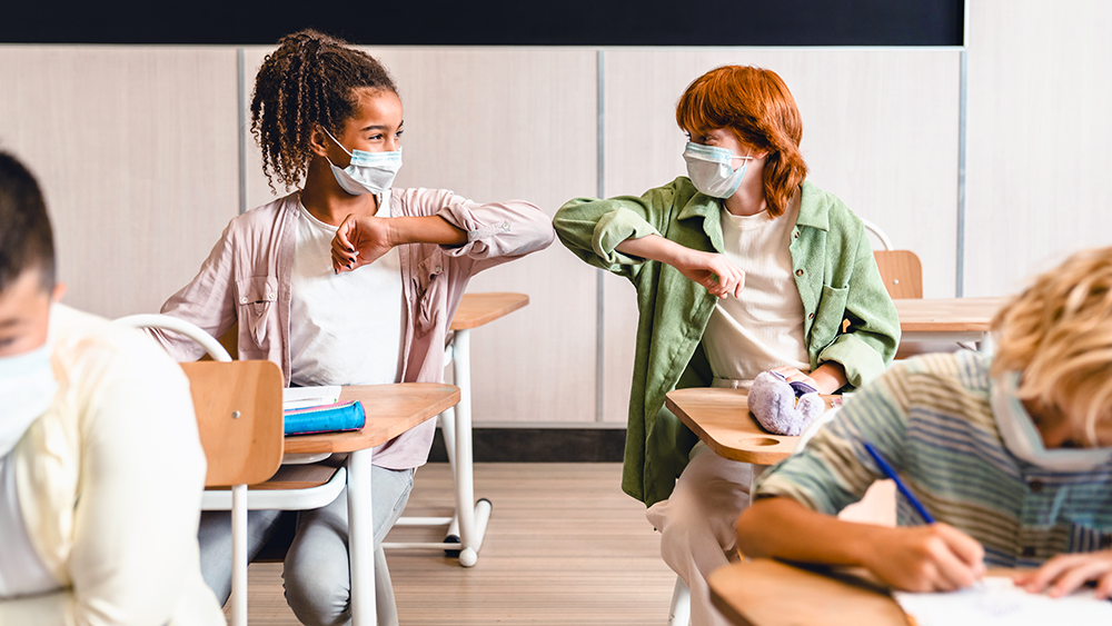 Zwei Mädchen im Schulzimmer tragen eine Maske und begrüssen sich mit dem Ellbogen