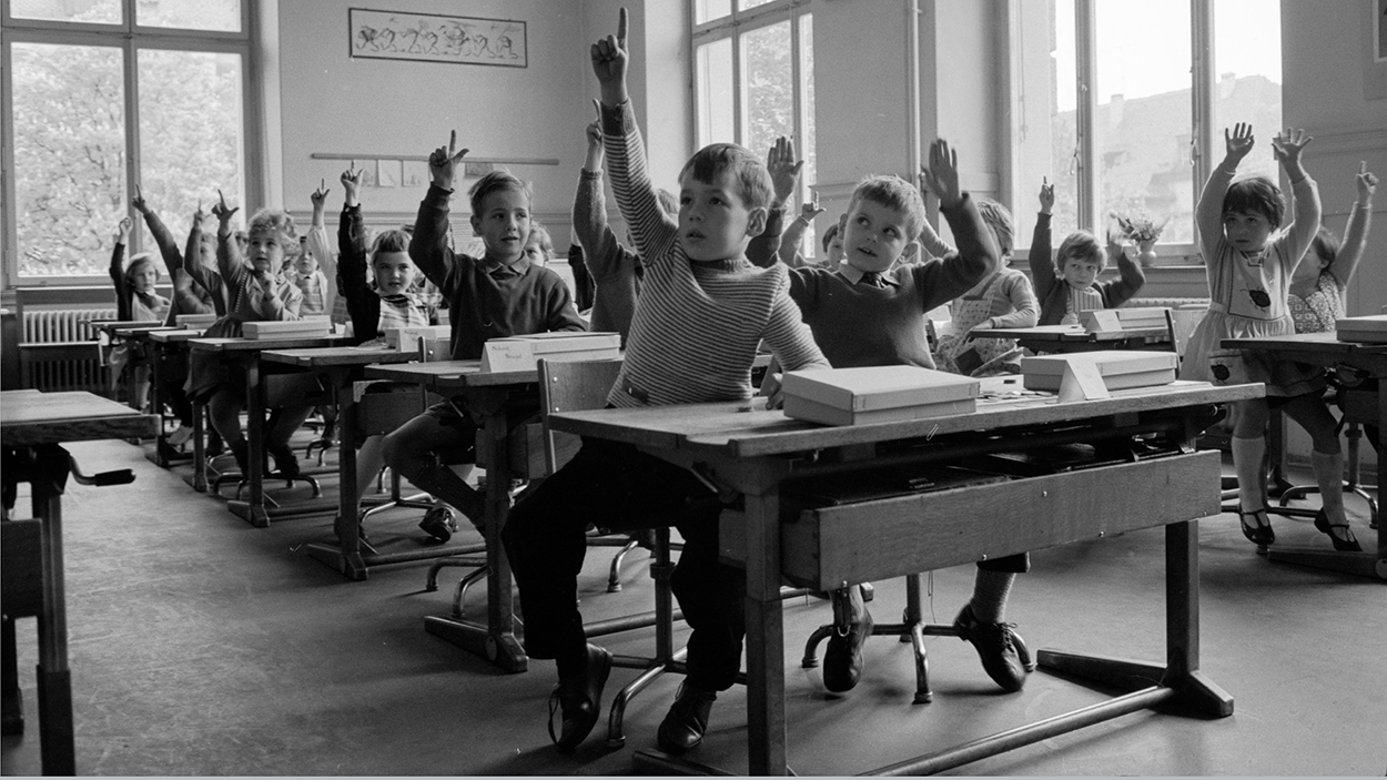 Schulkinder sitzen zu zweit in Schulbänken und strecken die Hand in die Luft