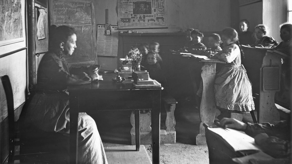 Vue sur une salle de classe vers 1920; une enseignante est assise au pupitre, on voit une douzaine d'élèves assis en rang