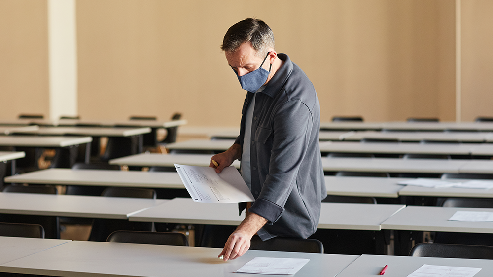 Ein Lehrer mit Maske verteilt Prüfungsmaterialien in einem noch leeren Prüfungsraum