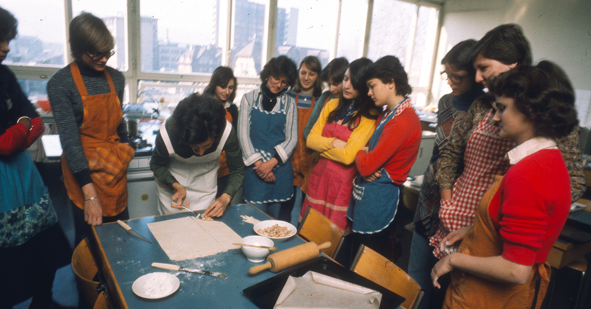 Mehrere junge Frauen stehen um einen Tisch und sehen einer Lehrerin beim Kochen zu